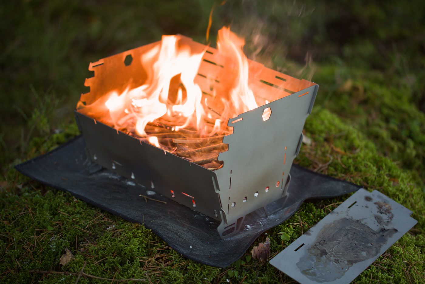 Titan1 titanium fire pit burning at dusk on its base mat, plancha plate alongside