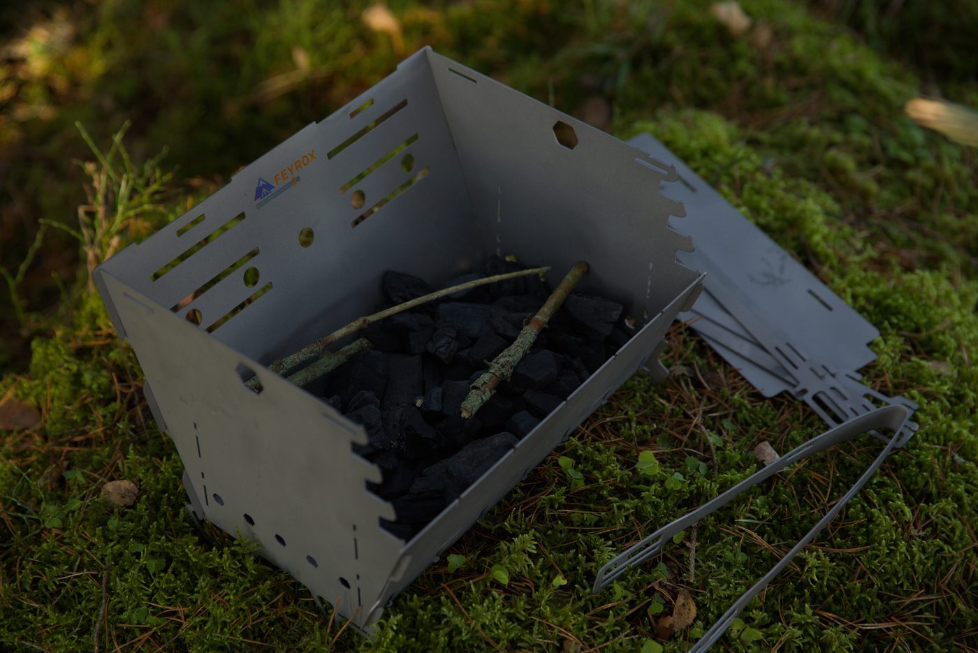 Titan1 titanium fire pit assembled on moss and loaded with charcoal, tongs and spare panels beside it